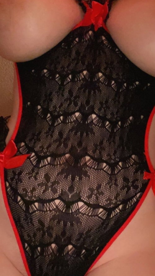 Close-up of a person wearing a black lace bodysuit with red trim and bows, showcasing a curvy figure in an indoor