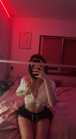 A woman with shoulder-length hair sits on a bed in a red-lit room, taking a mirror selfie in a tied white shirt and