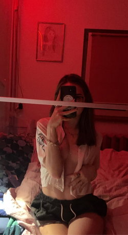 A woman with dark hair takes a mirror selfie in a red-lit room, wearing an unbuttoned white shirt and black shorts,