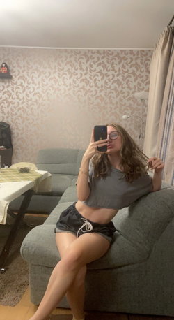 A woman with wavy hair and glasses sits on a gray couch, wearing a crop top and shorts, taking a mirror selfie in a