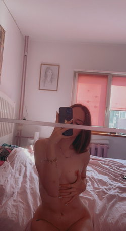 A woman with shoulder-length brown hair takes a mirror selfie in a bedroom, nude and covering her chest, with pink