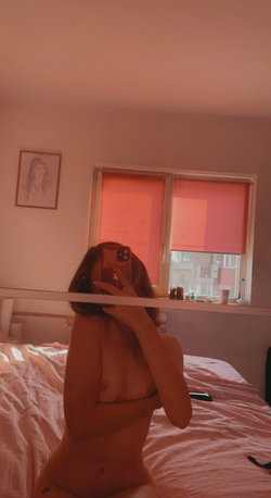 A woman with shoulder-length hair takes a mirror selfie in a bedroom, topless and sitting on a bed with pink bedding,