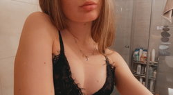 Blonde woman in black lace lingerie, wearing a delicate necklace, posing in a bathroom with tiled walls and shower