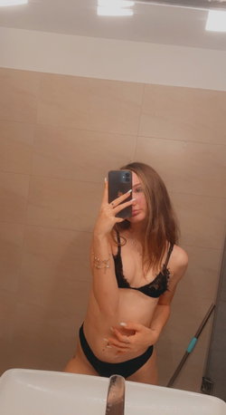A woman with long brown hair takes a mirror selfie in a bathroom, wearing a black lace bra and matching thong.