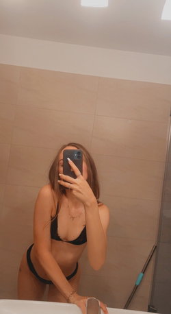 A woman with long brown hair takes a mirror selfie in a bathroom, wearing a black lace bra and matching thong.