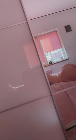 A person's bare back and shoulders are visible in a mirror reflection, lying on a bed in a pink-lit bedroom with a