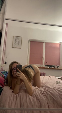 A woman with blonde hair and glasses takes a mirror selfie in bed, wearing gray thong underwear and showing a tattoo on