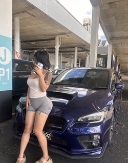Woman with tattoos in a white top and gray shorts leans on a blue car in a parking garage.