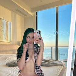 A woman with vibrant green hair takes a mirror selfie in a hotel room, topless and wearing a plaid shirt open over pink