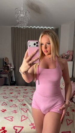 Curvy blonde woman in pink clothing taking a selfie in a bedroom.