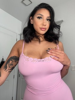 Curvy woman with dark hair in a pink lace-trimmed bodysuit, posing indoors with visible arm tattoo.