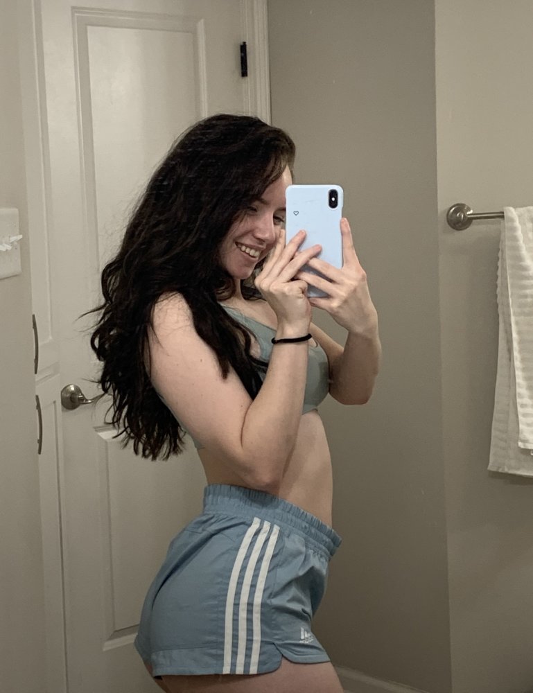 Brunette woman with long curly hair takes a mirror selfie in a bathroom, wearing a light blue sports bra and matching