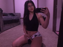 A young woman with long dark hair and glasses takes a mirror selfie in a bedroom, wearing a black crop top and