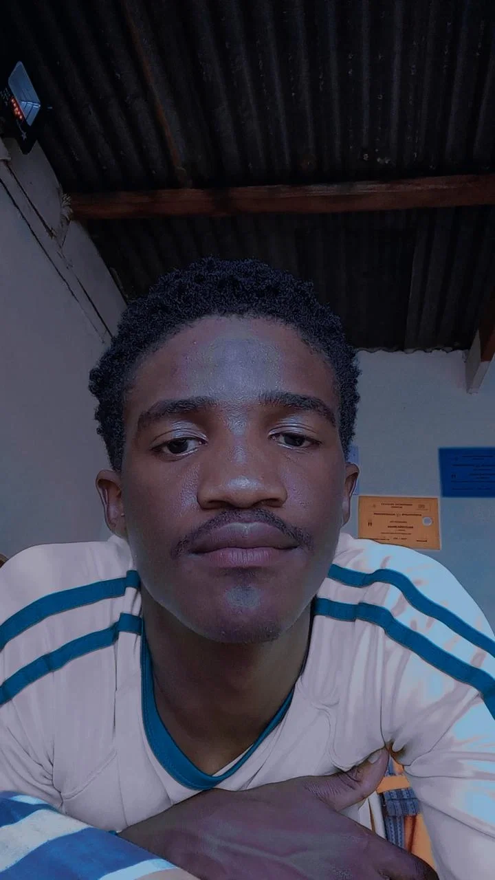 Close-up selfie of Black man with mustache wearing blue-striped shirt in room with metal roof.