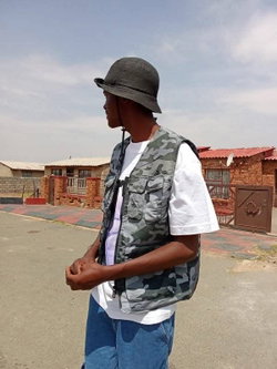 Side profile of dark-skinned man in black bucket hat, camo vest over white t-shirt, jeans, standing outdoors near brick