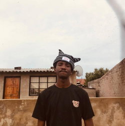 Man in black teddy bear t-shirt and black-white striped cat-ear beanie, smiling outdoors by brick house.