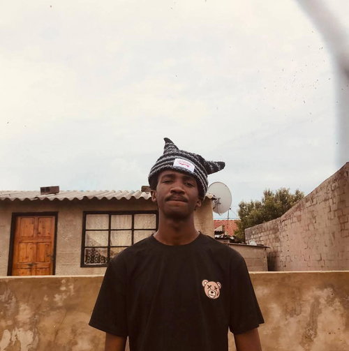 Man in black teddy bear t-shirt and black-white striped cat-ear beanie, smiling outdoors by brick house.