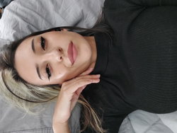 Woman with long dark hair and light-tipped ends lies on a bed with a neutral expression.