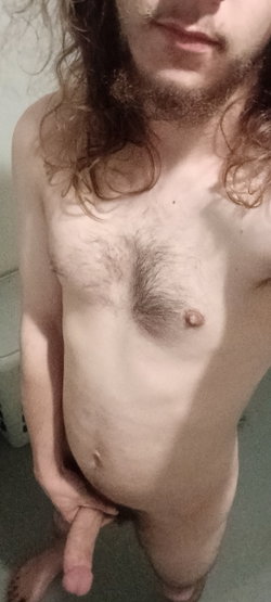 A person with curly hair and a beard poses topless in a close-up shot.
