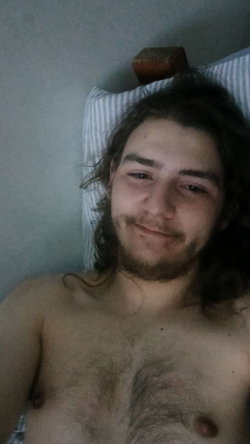 Shirtless man with long dark hair and beard lying in bed, smiling at the camera.
