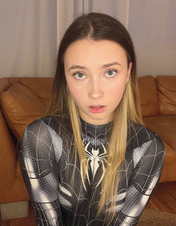 A young woman with long ombre hair and blue eyes wears a black and white spider-themed bodysuit, sitting on a brown