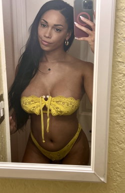 A woman with long dark hair takes a mirror selfie in a yellow lace lingerie set, showing a curvy figure in a bathroom