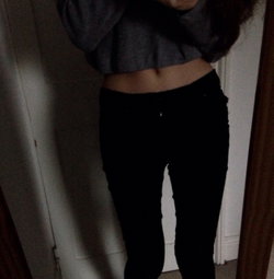 Person in a gray cropped sweatshirt and black pants, standing indoors with midriff exposed, likely taking a mirror