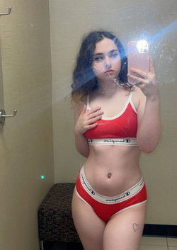 Curvy woman with dark curly hair takes mirror selfie in red Champion lingerie, hand on chest, small heart tattoo