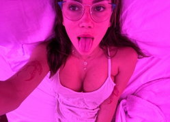 Woman with glasses and tattoos lies in bed under pink lighting, wearing a white tank top and sticking out her tongue.