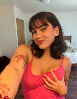 A woman with shoulder-length dark hair and bangs smiles in a bedroom selfie, wearing a pink tank top and showing arm