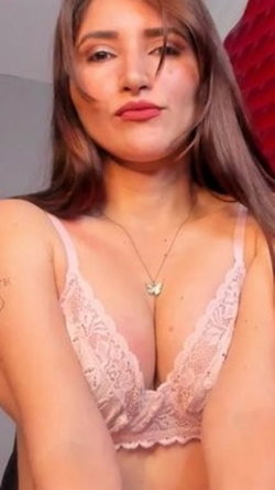 A woman with long brown hair wears a light pink lace bra, posing indoors with a red pillow behind her.