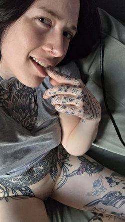 Tattooed woman with dark hair smiles while lying on a couch in a grey top.