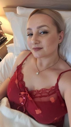 Blonde woman in red lace lingerie lying in bed, smiling softly at the camera with makeup and a delicate necklace.