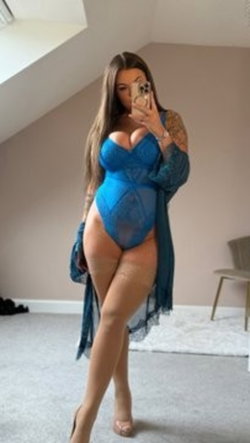 Curvy woman with long dark hair takes mirror selfie in blue lace bodysuit and sheer robe, standing in a softly lit room.
