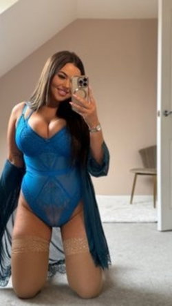 Brunette woman in blue lace bodysuit and thigh-high stockings kneels on carpet, holding phone for mirror selfie in