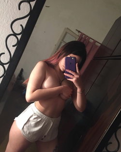 A woman with pink hair takes a selfie in a mirror wearing white shorts.