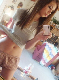 A young woman with long brown hair takes a mirror selfie in a gray crop top and plaid shorts, showing her midriff in a