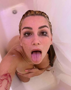 A woman with wet hair and makeup in a bathtub, sticking her tongue out, showing a tattoo on her arm.