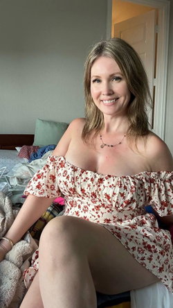 A smiling woman with shoulder-length light brown hair sits on a bed wearing an off-the-shoulder floral dress, in a