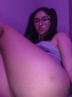 A woman with dark hair and glasses poses in a purple-lit room, wearing a strapless top, with one leg raised toward the