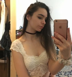 Slim woman with long brown hair takes a selfie in a white lace top and choker.