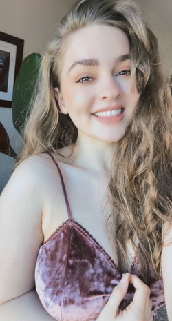 Blonde woman with wavy hair smiling in a purple velvet top, indoors with a plant and framed art in the background.