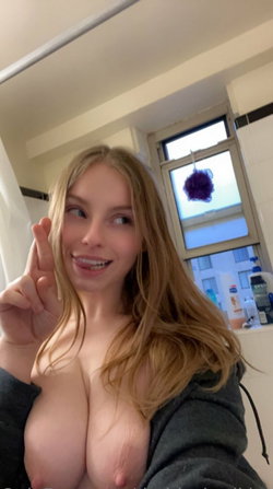 Blonde woman with bare breasts in a bathroom, wearing an open hoodie and smiling at the camera.