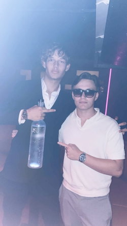 Two men posing in neon-lit club, one in black suit jacket and sunglasses holding water bottle, other in white shirt and