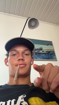 Man in backwards cap and black t-shirt holding joint in mouth, pointing at camera in room with lamp and boat painting.