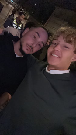 Two smiling young men in casual clothes take a selfie outdoors at night near police officers.