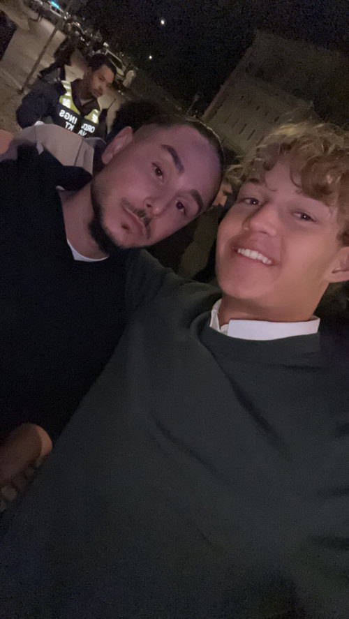 Two smiling young men in casual clothes take a selfie outdoors at night near police officers.