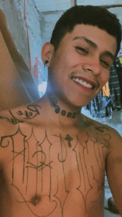 A smiling man with tattoos on his chest and neck stands in a dimly lit room.