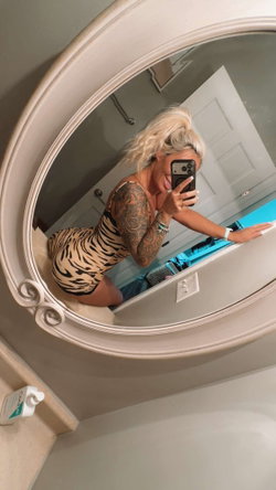Blonde woman with full sleeve tattoos takes mirror selfie in a tiger-print bodysuit, smiling in a bathroom setting.