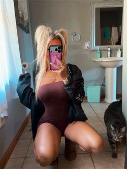 Blonde woman in a maroon bodysuit and black jacket takes a mirror selfie in a bathroom, kneeling with a cat nearby.